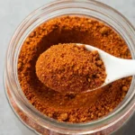 Homemade chili seasoning recipe in jar with chili powder, cumin, and smoked paprika spices, measuring spoons, and dried chilies on wooden background
